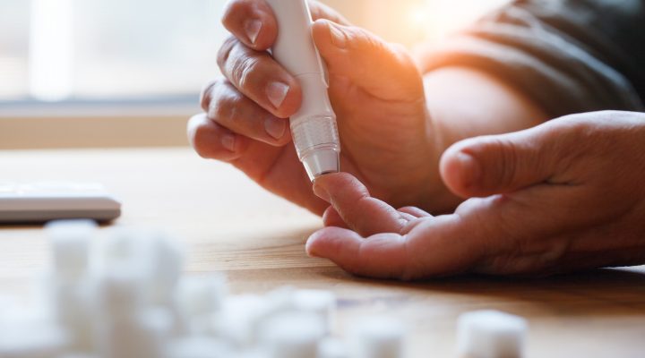 woman hands using lancet on finger at home to check blood sugar level, glucometer and sugar cubes on wooden table close up, diabetes concept, elderly health care, sunny morning