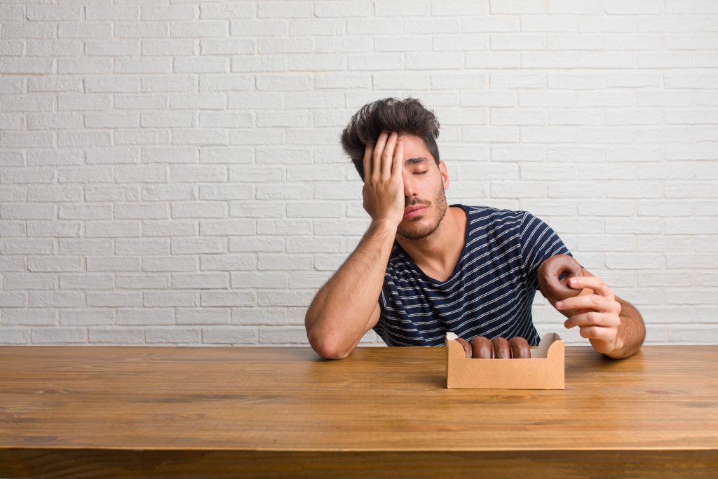 A man looking stressed and tired eating a box of chocolate donuts